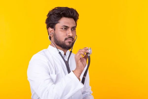 Male Indian doctor holding a stethoscope in his hands to listen to a patient posing on a yellow background. Concept of treatment and recovery. Copyspace. — Stock Photo, Image