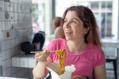 Bir kafede udon erişte yiyen ve pencereden dışarı bakan genç neşeli bir kız portresi. Sağlıklı Asya mutfağı kavramı.