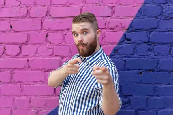 Divertido joven tipo elegante con un bigote y una barba en una camisa elegante posando en una pared de ladrillo azul-púrpura y señalando los dedos en la cámara. Concepto de promociones y descuentos . — Foto de Stock