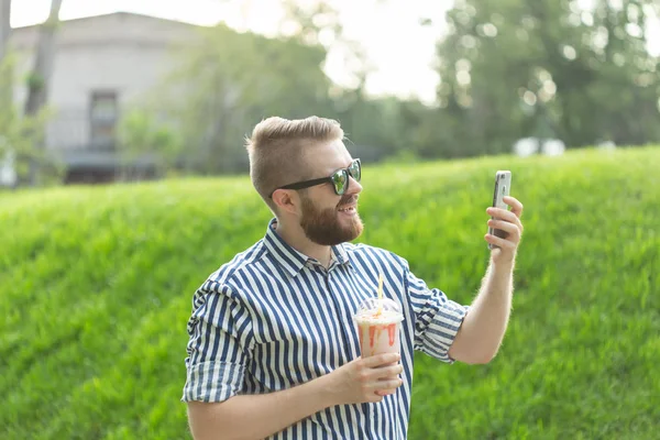 Vista lateral de un joven elegante con gafas y una barba con un batido en las manos hablando en video con sus amigos usando un teléfono inteligente e Internet gratuito de alta velocidad en el parque en un cálido día de verano — Foto de Stock