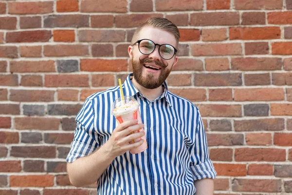 Joven chico hipster positivo en gafas y con barba está sosteniendo un cóctel con una pipa posando contra una pared de ladrillo. El concepto de vacaciones y vacaciones de verano . — Foto de Stock