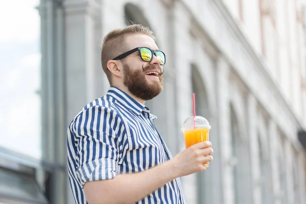 Vista lateral de un joven empresario bastante alegre con un bigote elegante y una barba con jugo en las manos, caminando por la ciudad después de un día de trabajo. Concepto de positivo y descanso . — Foto de Stock