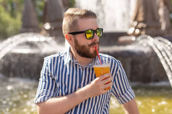 Lindo hipster joven y elegante con bigote y barba y gafas bebiendo jugo con una pajita mientras está sentado en un parque de la ciudad sobre el fondo de una fuente. Concepto vacaciones de verano . — Foto de Stock