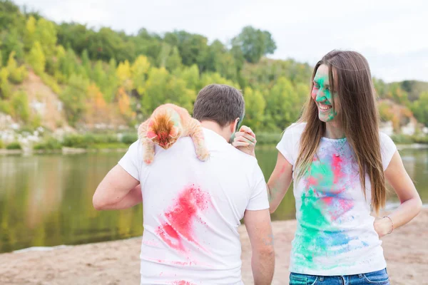 Festival holi, férias, turismo e conceito de natureza - Casal vestido com camisas brancas segurando gato e coberto com poeira colorida — Fotografia de Stock