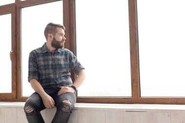 Şık giysiler ile sevimli genç hipster adam büyük pencerelerin yakınında bir akıllı telefon ve kablosuz internet kullanarak sosyal ağlarda oturuyor. İnternet bağımlılığı kavramı.