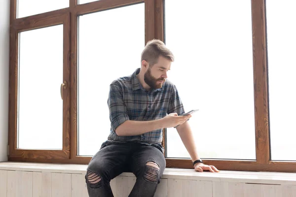 Cute young hipster guy with stylish clothes is sitting on social networks using a smartphone and wireless internet near large windows. Concept of internet addiction.