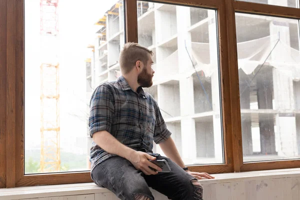 Młody przystojny człowiek Hipster siedzi na parapecie w pobliżu dużego okna z smartphone w dłoniach i patrząc na budowie w domu. Koncepcja początkującego architekta. — Zdjęcie stockowe