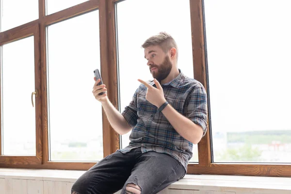Lindo chico positivo joven hipster con una barba está sentado en el alféizar de la ventana grande y hablando en la comunicación de vídeo utilizando un teléfono inteligente e Internet inalámbrico de alta velocidad . — Foto de Stock