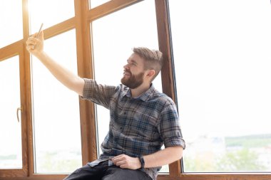 Olumlu yakışıklı hipster adam yan görünümü ve büyük bir pencere tarafından bir pencere üzerinde otururken bir akıllı telefonda selfie alarak. Gadget'lar ve modern teknolojiler kavramı.