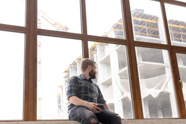 Genç yakışıklı adam hipster elinde bir akıllı telefon ile büyük bir pencerenin yakınında pencere üzerinde oturan ve evde şantiye bakıyor. Acemi mimar konsepti.