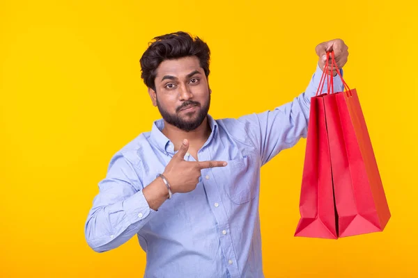 Örömteli indiai férfi tartja piros bevásárló táskák, sárga háttérrel. A koncepció a vásárlás a szupermarketben és ajándékokat. Reklámfelület. — Stock Fotó