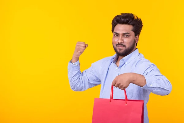 Fiatal csinos indiai férfi egy szakállas tart piros bevásárló táskák pózol a sárga háttér másolási tér. Szupermarket vásárlási koncepció. — Stock Fotó