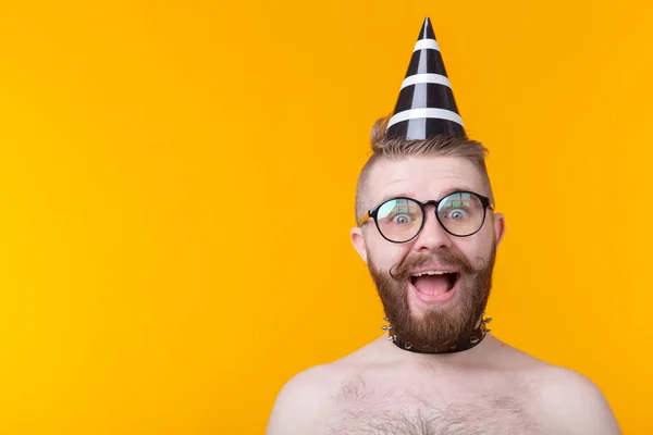 Crazy cheerful hipster guy in a paper cap with a mustache and beard screaming for joy posing shirtless with a leather collar on a yellow background with copy space