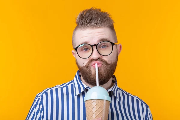 Leuke grappige jonge man met een snor en baard drinken van een cocktail met een rietje op een gele achtergrond. Het concept van zomerse desserts en drankjes. — Stockfoto