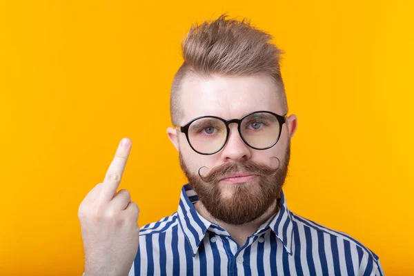 Ein wütender junger Mann mit Brille, Schnurrbart und Bart zeigt den Mittelfinger, der auf gelbem Grund posiert. Konzept der Verleugnung und Verärgerung. — Stockfoto