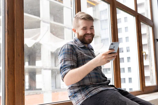 Lindo chico hipster joven con ropa elegante está sentado en las redes sociales utilizando un teléfono inteligente e Internet inalámbrico cerca de grandes ventanas. Concepto de adicción a Internet . — Foto de Stock