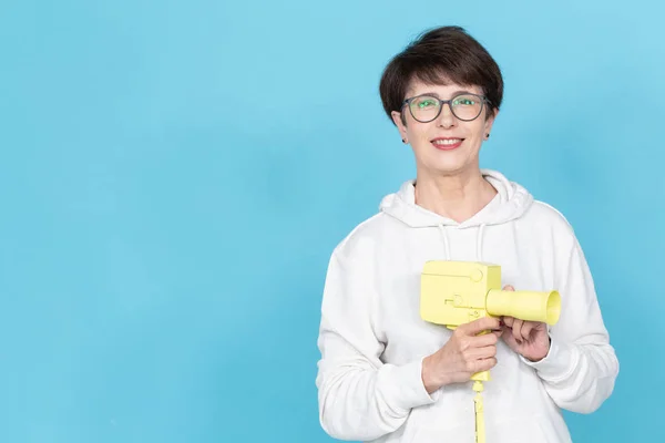 Mujer de mediana edad con un corte de pelo corto en un suéter y gafas que sostienen una cámara de cine vintage amarilla sobre fondo azul con espacio para copiar. Concepto de grabación de vídeo — Foto de Stock