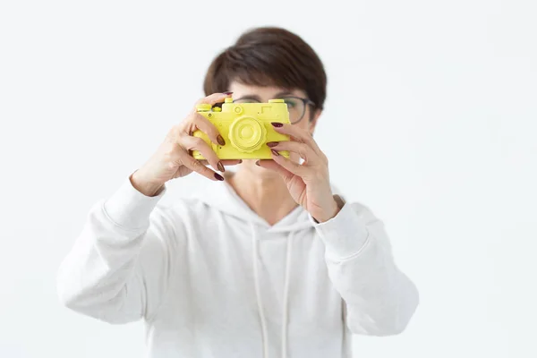 Positive middle-aged woman in glasses holds in hand a vintage yellow camera posing on a white background. Concept of photo lovers and hobbies — Stock Photo, Image