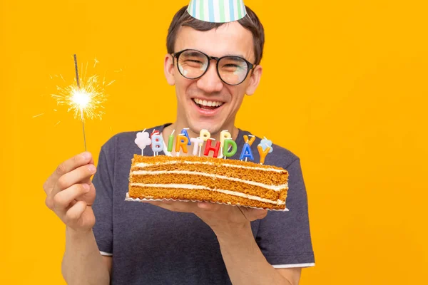 Pazzo allegro giovane ragazzo asiatico con gli occhiali che tiene una candela accesa tra le mani e una torta fatta in casa di congratulazioni su uno sfondo giallo. Concetto di compleanno e anniversario . — Foto Stock