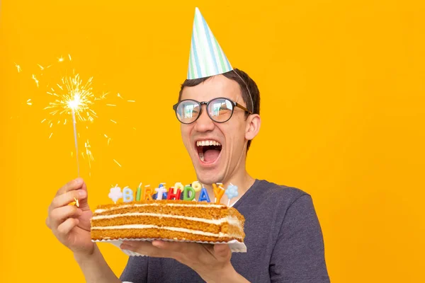 Divertente ragazzo positivo in occhiali tiene tra le mani una torta fatta in casa con l'iscrizione buon compleanno in posa su uno sfondo giallo. Concetto di vacanze e anniversari . — Foto Stock