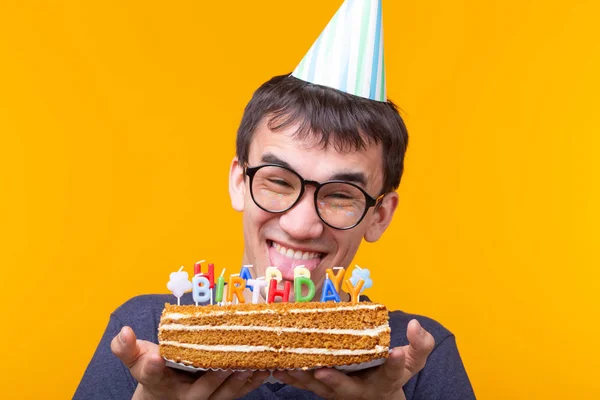 Ritratto di un simpatico ragazzo positivo con un tappo di carta e occhiali che tiene tra le mani una torta fatta in casa di congratulazioni su uno sfondo giallo. Concetto e divertimento e celebrazione . — Foto Stock
