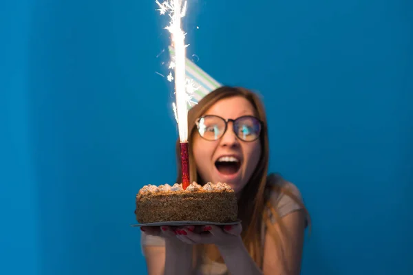 Jovem estudante menina borrada alegre em óculos segurando um bolo de parabéns com uma vela em pé sobre um fundo azul. Conceito de aniversário . — Fotografia de Stock