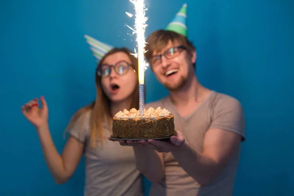 Веселая молодая пара очаровательный парень и милая девушка в бумажных шляпах делают глупое лицо и держать в руках торт день рождения стоя на синем фоне . — стоковое фото