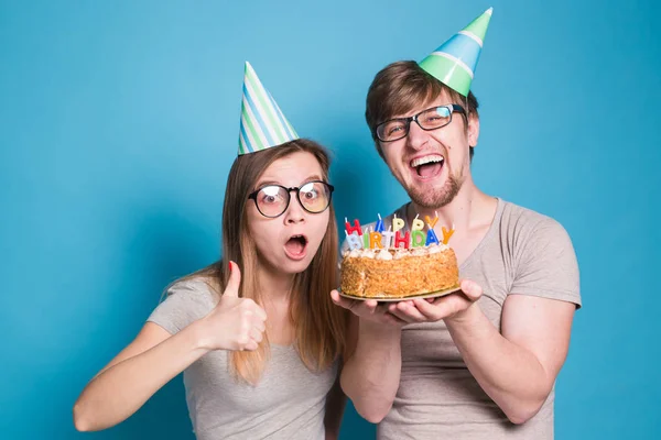 명랑한 젊은 부부 매력적인 남자와 종이 모자에 귀여운 소녀는 어리석은 얼굴을하고 파란색 배경에 서 생일 케이크를 손에. — 스톡 사진