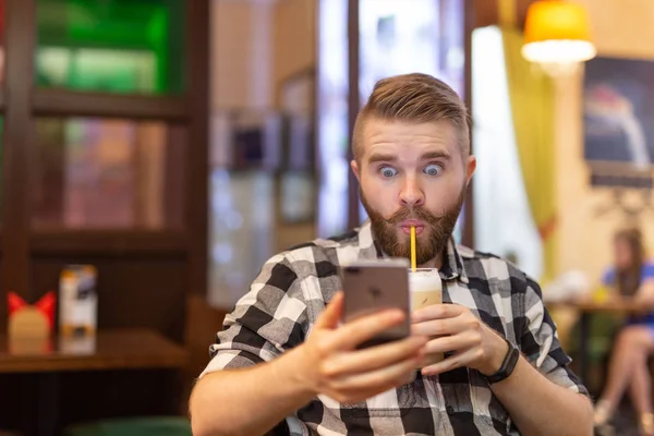 Sorprendido joven hipster bebiendo cacao de una paja y mirando los mensajes en las redes sociales mientras estaba sentado en una mesa en un café. El concepto de chat y redes sociales . — Foto de Stock