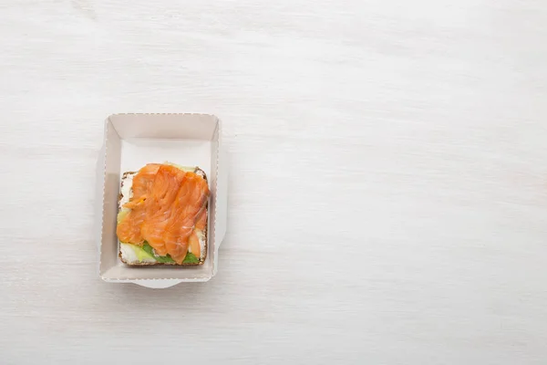 Στο κουτί του γεύματος δίπλα στα χόρτα και τις ντομάτες σε λευκό φόντο με φωτοτυπία, υπάρχει ένα σάντουιτς με απαλά τυριά και κόκκινα ψάρια. Έννοια ενός υγιεινού σνακ. — Φωτογραφία Αρχείου