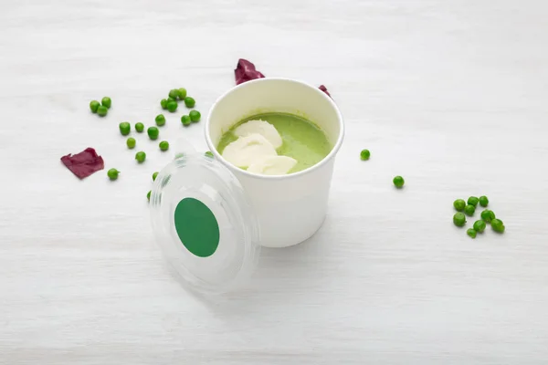 Чашка зеленого супа с кусочками овощей лежит на белом столе с зеленым горошком. Концепция здорового питания — стоковое фото