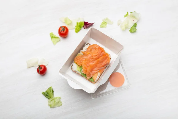 Στο κουτί του γεύματος δίπλα στα χόρτα και τις ντομάτες σε ένα λευκό τραπέζι, θα δείτε ένα σάντουιτς με απαλό τυρί και κόκκινα ψάρια. Έννοια ενός υγιεινού σνακ. — Φωτογραφία Αρχείου