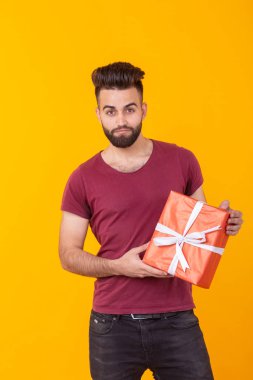 Sarı bir arka plan üzerinde poz ve kırmızı bir hediye kutusu tutan sakallı yakışıklı bir genç erkek hipster Portresi. Hediye ve indirim kavramı.