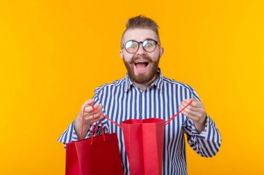 Gözlük ve sakal ile sürpriz neşeli genç yakışıklı adam sarı bir arka planda ayakta, alışveriş için paketler içine bakıyor. Başarılı satın alma kavramı. Reklam alanı.