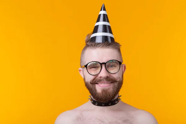 Loco chico hipster alegre en una gorra de papel con un bigote y barba gritando de alegría posando sin camisa con un collar de cuero sobre un fondo amarillo con espacio para copiar — Foto de Stock