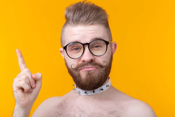 Funny young man with a mustache and a beard without a shirt and glasses shows on a yellow background. Concept of non-standard advertising.