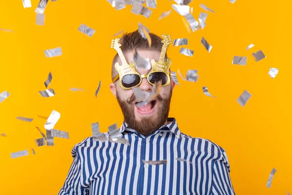 Joven hipster alegre macho con una gorra de papel y gafas se ríe felizmente entre confeti volador sobre un fondo amarillo. El concepto de una mega fiesta y vacaciones . — Foto de Stock