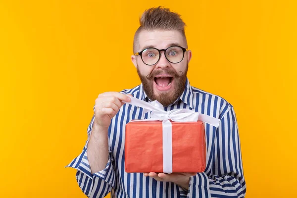 Surprised young hipster man with mustache and beard in surprise unpacks a red box with gifts on a yellow background, close up. The concept of gifts and surprises for the holiday