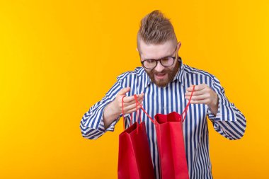 Gözlük ve sakal ile sürpriz neşeli genç yakışıklı adam sarı bir arka planda ayakta, alışveriş için paketler içine bakıyor. Başarılı satın alma kavramı. Reklam alanı.