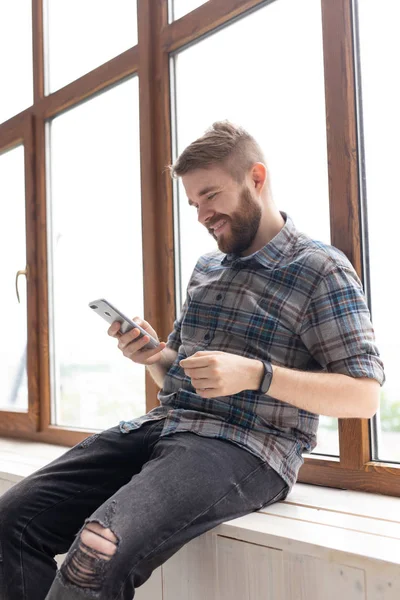 Lindo chico hipster joven con ropa elegante está sentado en las redes sociales utilizando un teléfono inteligente e Internet inalámbrico cerca de grandes ventanas. Concepto de adicción a Internet . — Foto de Stock
