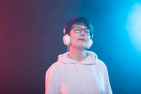 Beautiful middle-aged woman in a white sweater and glasses is listening to music while posing on a red-blue background in a studio. The concept of a favorite hobby. Advertising space. — Stock Photo, Image