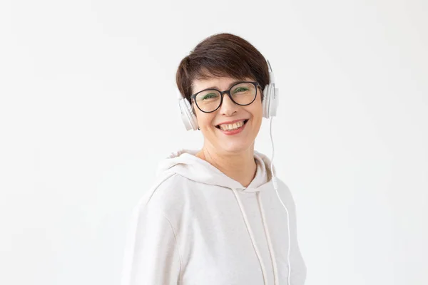 Cute positive middle-aged woman in sweater and glasses is listening to music with wire headphones standing on a white background with copy space. Concept of hobbies and subscriptions to favorite radio — Stock Photo, Image