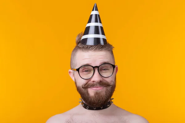 Loco chico hipster alegre en una gorra de papel con un bigote y barba gritando de alegría posando sin camisa con un collar de cuero sobre un fondo amarillo con espacio para copiar — Foto de Stock