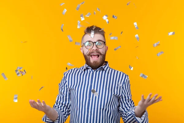 Crazy young positive hipster guy with a beard laughs happily among flying kofetti on a yellow background. The concept of holidays and sales