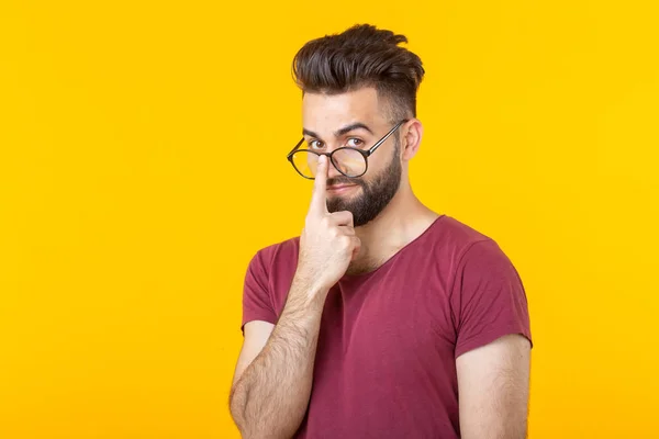 Joven hombre hipster pensativo con una barba sosteniendo sus gafas mientras está de pie sobre un fondo amarillo. El concepto de atención y elección difícil — Foto de Stock
