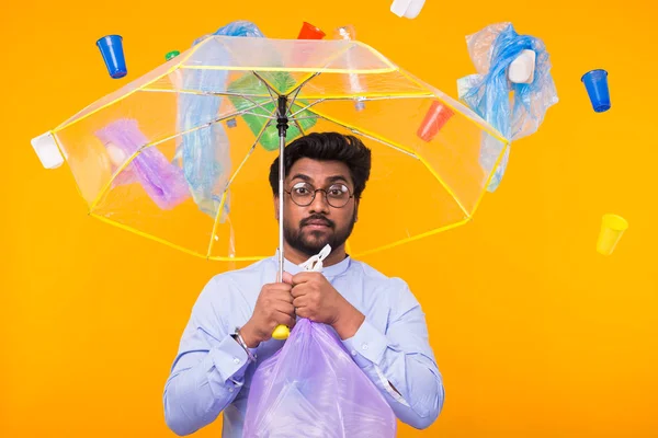 Dia Mundial do Meio Ambiente, problema de reciclagem de plástico e conceito de desastre ambiental - surpreendeu o homem indiano escondendo-se do lixo sob um guarda-chuva no fundo amarelo — Fotografia de Stock