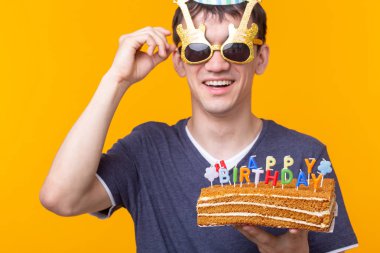 Elinde yanan bir mum tutan gözlük ve sarı bir arka plan üzerinde bir tebrik ev yapımı pasta ile Crazy neşeli genç Asya adam. Doğum günü ve yıldönümü kutlama konsepti.