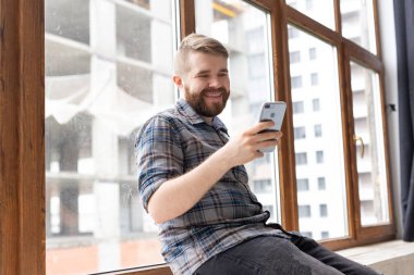 Bir sakal ile sevimli genç pozitif adam hipster büyük pencere tarafından pencere üzerinde oturuyor ve bir akıllı telefon ve kablosuz yüksek hızlı Internet kullanarak video iletişimi üzerinde konuşuyor.