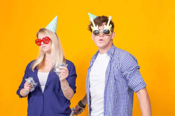 Young cheerful positive couple of funny glasses and a paper cap rejoice and shout on a yellow background with flying confetti. Conception of a holiday and fun. — Stock Photo, Image