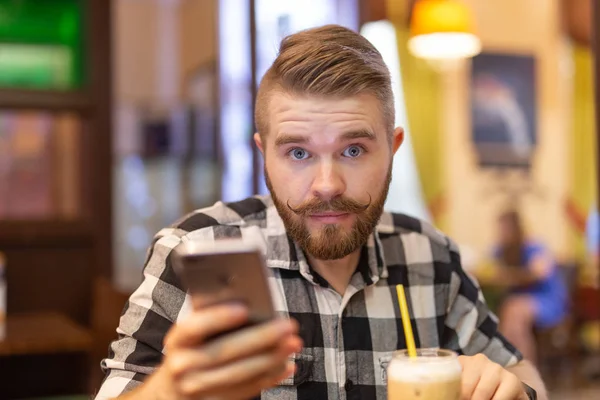 Pensativo joven y elegante joven hipster hombre con bigote y barba está buscando en la red social en el teléfono inteligente utilizando Internet de alta velocidad gratuita. Concepto de comunicación en las redes sociales . — Foto de Stock